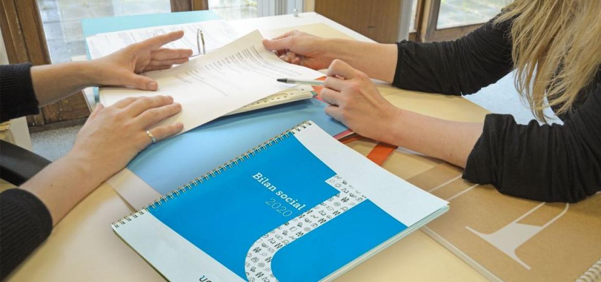 deux personnes assises de part et d'autre d'un bureau. L'une des deux pointe des informations écrites sur une feuille de papier.